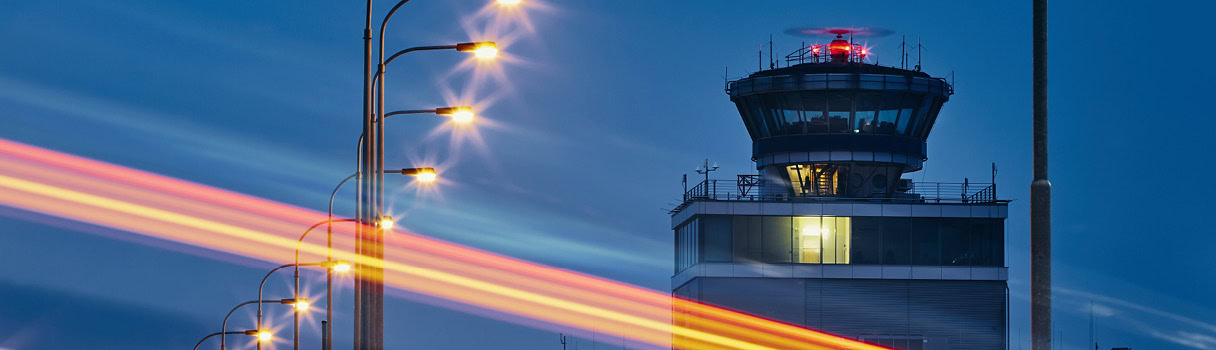 Torre de control del aeropuerto de noche con franjas luminosas de los vehículos que pasan, simbolizando la sincronización horaria.