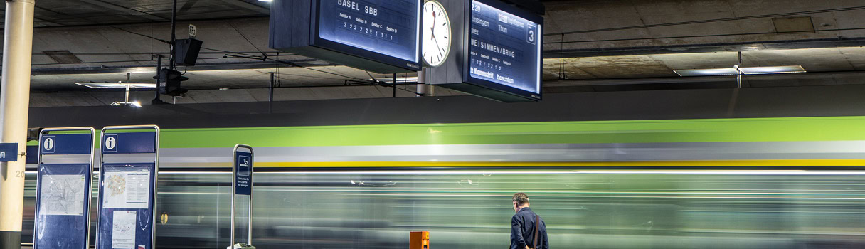 A picture of a railway station with a train passing by. In the picture, there is also a swiss railway clock by Mobatime.