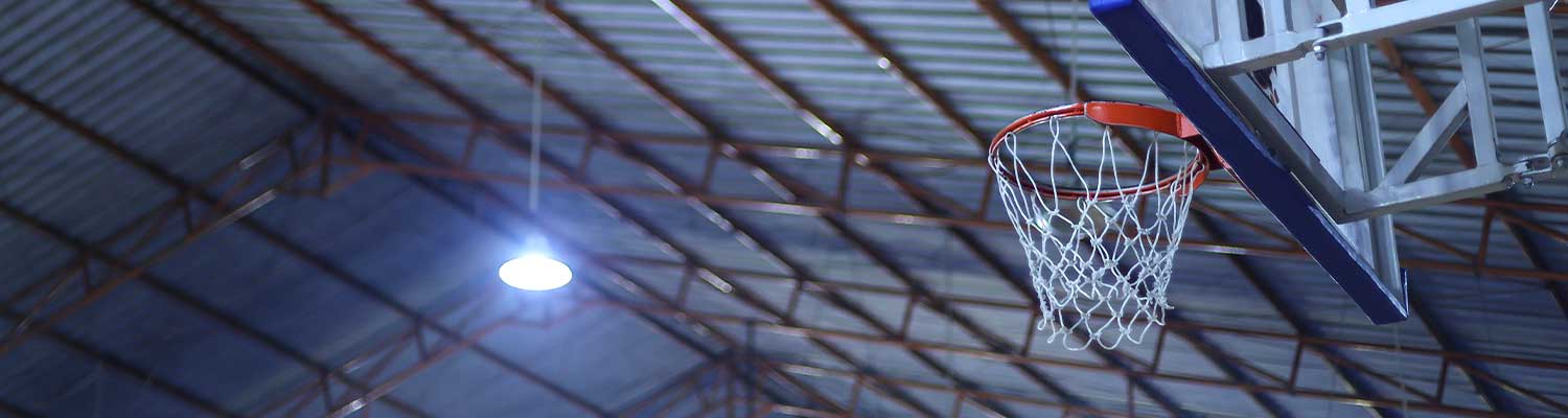 Indoor basketball hoop mounted to a metal backboard, set against the high ceiling of a sports and leisure facility with exposed beams and bright overhead lighting
