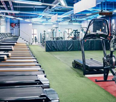 Modern indoor gym with rows of treadmills, exercise machines, and a prominent digital clock displaying 10:08, set against a clean, well-lit space with blue ceiling lights.