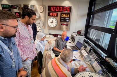 Des ingénieurs et des représentants du projet observent les tests en direct des horloges maître et esclave lors du test de réception en usine (FAT) pour le projet de train à grande vitesse égyptien au siège de Moser-Baer, avec des affichages synchronisés de l'heure visibles à l'arrière-plan.